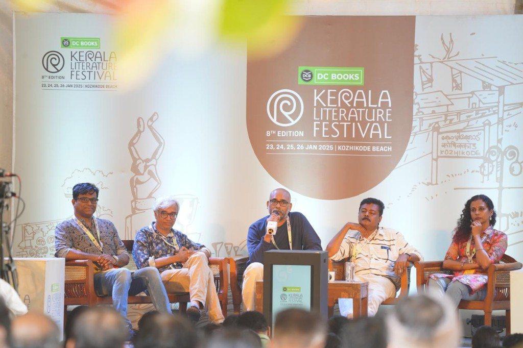 O.P. Suresh speaking on a Kerala Literature Festival panel. The backdrop reads 8th edition, 23, 24, 25, 26 January 2025, Kozhikode Beach.