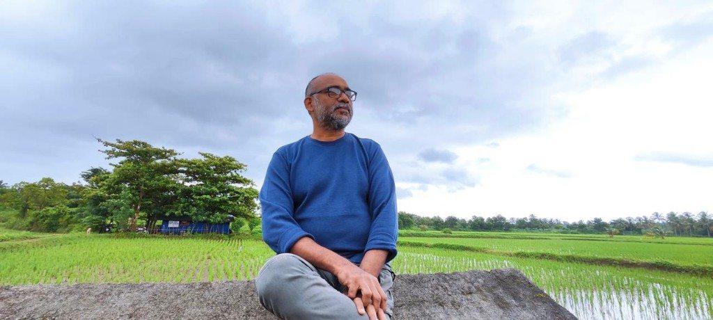 O.P. Suresh seated on a rock above a green paddy field under cloud.